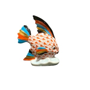 May include: A ceramic fish figurine with a white body, orange scale pattern, and colorful fins. The fins feature blue, orange, yellow, and black accents. The fish rests on a white cloud-shaped base, creating a decorative nautical theme.