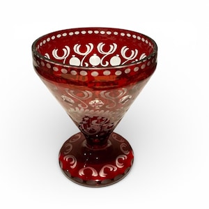 May include: A ruby red glass goblet with a flared bowl and a round base. The goblet is decorated with white etched designs, including dots and swirling patterns. The rim and base have a decorative border.