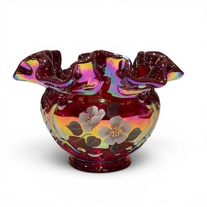 Könnte beinhalten: Eine tiefrote, irisierende Glasschale mit einem gerüschten, gewellten Rand und einem handgemalten Blumenmuster aus weißen Blütenblättern und grünen Blättern. Die Glasoberfläche zeigt einen mehrfarbigen metallischen Glanz auf einem runden Fuß.