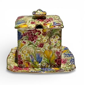 May include: A floral-patterned ceramic sugar bowl with a matching lid and a small, attached saucer. The design features a mix of colorful flowers, including purple, yellow, and blue blooms, set against a cream background. The edges are trimmed with gold.