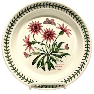 May include: A round, off-white plate with a floral design. It features red and pink Gazania flowers and butterflies. The plate has a green leaf border and the text "GAZANIA RINGENS Treasure Flower".