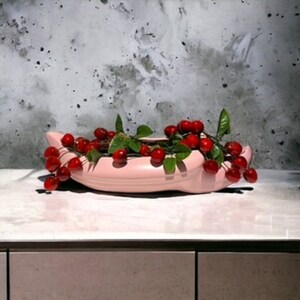 May include: A pink ceramic banana-shaped bowl filled with red cherries and green leaves.
