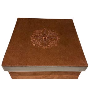 Peut inclure: Une boîte carrée marron avec un motif brodé sur le couvercle. La boîte a une texture semblable à du daim et un bord marron plus foncé. Le motif brodé est un motif symétrique détaillé dans une teinte plus claire de marron.