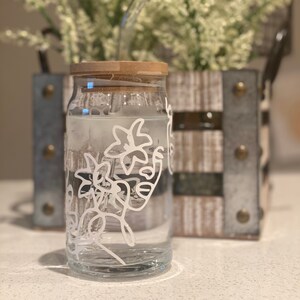 May include: A clear glass jar with a wooden lid and a white floral design. The jar is filled with water and has a straw in it.