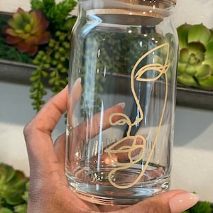 May include: A clear glass jar with a bamboo lid and a gold line drawing of a face on the front. The jar is being held by a hand.