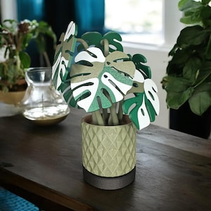 May include: A decorative monstera plant in a light green geometric patterned pot. The plant features large, stylized leaves in shades of green, white, and gold glitter. The pot sits on a dark brown base, creating a modern, artistic display.