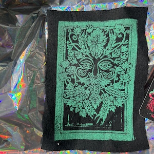 May include: A green and black fabric patch with a green, stylized image of a face surrounded by leaves. The face has large eyes and a beard. The image is framed by a black border.