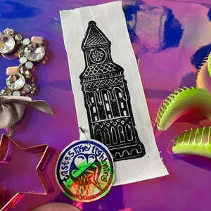 May include: A black and white print of a clock tower on a white fabric scrap. A circular tag with the text "tastes like lightning" and a colorful graphic is attached. Other jewelry and a Venus flytrap are also visible.