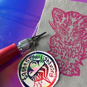 Pode incluir: Uma ferramenta de pirogravura com cabo rosa e ponta prateada está posicionada perto de uma amostra de tecido cinza com um desenho de animal rosa. Uma etiqueta redonda com o texto "tastes like lightning" e um gráfico de raio também está presente.