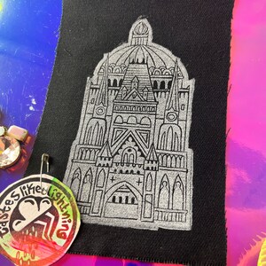 May include: Black fabric patch featuring a detailed silver illustration of a cathedral. The patch is secured with a safety pin. A circular sticker with the text "babes like lightning" is also visible.