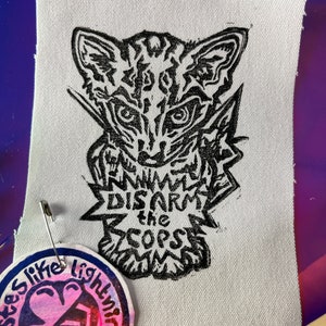 Pode incluir: Um patch de tecido branco com uma impressão em linóleo preto de um gato estilizado com o texto "DISARM the COPS". Uma etiqueta redonda com o texto "tastes like lightning" está presa ao patch.
