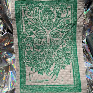 Op de afbeelding: Groene en witte stoffen patch met een groen bladerontwerp dat een gezicht voorstelt. Het gezicht heeft twee ogen en een mond. Het ontwerp is omlijst door een groene rand.