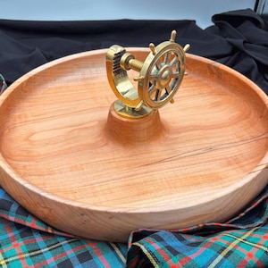 May include: A round wooden serving tray with a nautical-themed brass ship's wheel centerpiece. The tray is made of light-colored wood and has a smooth finish. The ship's wheel is detailed and adds a decorative touch.