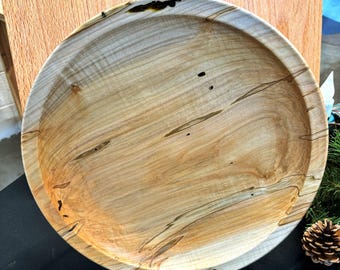 May include: A round, wooden serving tray with a natural wood grain pattern. The tray has a light brown color with darker streaks and knots. The edges are slightly raised. The tray is displayed on a black surface.
