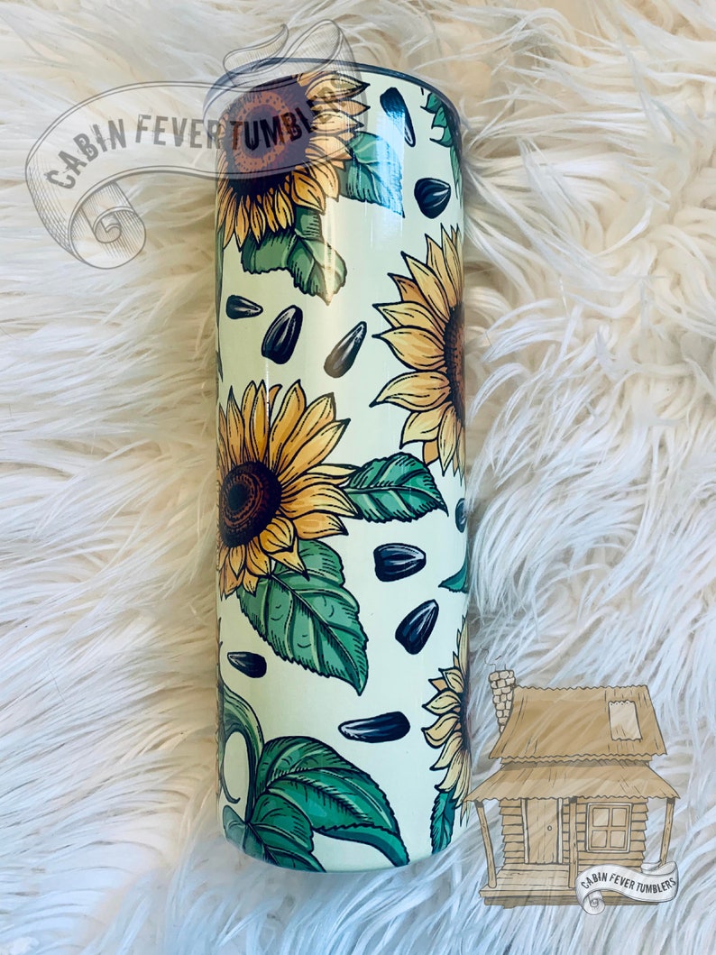 May include: A stainless steel tumbler with a white background and a pattern of sunflowers, leaves, and sunflower seeds. The tumbler is 20 ounces and has a double-wall insulated design. The text "CABIN FEVER TUMBLERS" is visible on the top of the tumbler.