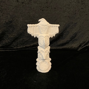 May include: White ceramic totem pole with an eagle at the top and a human face in the middle. The pole is decorated with carvings of feathers and other designs.