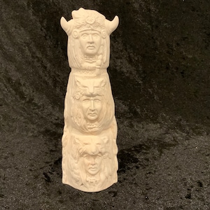 May include: White ceramic totem pole with three carved faces. The top face has horns and a headdress. The middle face has a headdress with feathers. The bottom face has a headdress with a lion's mane.
