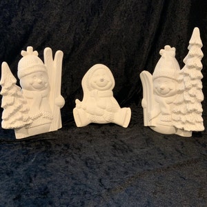 Puede incluir: Tres figuras de muñeco de nieve de cerámica blanca. Un muñeco de nieve sostiene esquís, otro está sentado con una bufanda y el tercero está de pie junto a un pequeño pino.