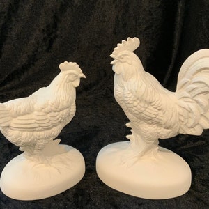 May include: Two ceramic figurines of a rooster and a hen. The rooster is standing with its tail feathers spread out. The hen is sitting with its head turned to the side. Both figurines are white and have a detailed, textured surface.