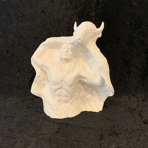 Op de afbeelding: Witte keramische sculptuur van een indiaanse man die een buffelhuid vasthoudt. De man draagt een ketting en heeft lang haar. De buffel is gedeeltelijk zichtbaar achter de man.