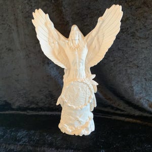 May include: White ceramic figurine of a Native American man with outstretched arms and eagle wings. The figurine is standing on a base with a detailed design.
