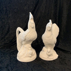 May include: Two white ceramic rooster figurines. One rooster is standing with a chick, the other is standing with its head held high.