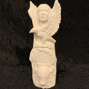 May include: White ceramic figurine of a Native American woman with an eagle perched on her shoulder. The woman is wearing a headdress and the eagle has its wings spread. The base of the figurine is decorated with a buffalo head and other Native American symbols.
