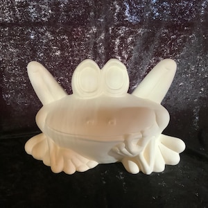 May include: White ceramic frog figurine with large eyes and a wide mouth. The frog is sitting on its hind legs with its front legs crossed.