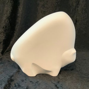 May include: White ceramic sculpture of a stylized animal, possibly a bison or bull, with a rounded, abstract form. The sculpture is positioned on a black surface.