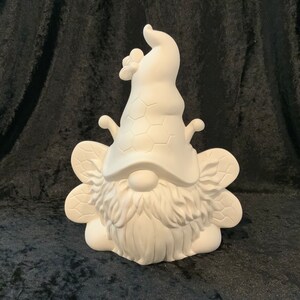 May include: White ceramic gnome figurine with a bee-themed hat and wings. The gnome has a long beard and a honeycomb pattern on its hat.