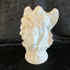 May include: A white ceramic sculpture featuring a woman with angel wings and long hair, standing beside a bison. The sculpture has a detailed design and is set on a round base. The woman is wearing a long dress and holding a feather.