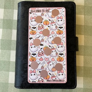 May include: A pink and white Halloween-themed scratch-off savings tracker featuring ghost and pumpkin illustrations. The tracker is placed on a black leather-bound planner. The text "Save $100 (5-25)" is visible at the top. The bottom of the tracker reads "Mara's Money Moves".