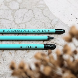 Op de afbeelding: Twee lichtblauwe potloden met zwarte tekst. Het bovenste potlood zegt "DU KANNST ALLES SCHAFFEN" met een ster aan elke kant. Het onderste potlood zegt "LEON" met een ster aan elke kant.