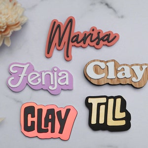 Könnte beinhalten: Sechs personalisierte Namensschilder mit verschiedenen Farben und Schriftarten. Die Namen sind "Manisa", "Fenja", "Clay", "Clay" und "Til".