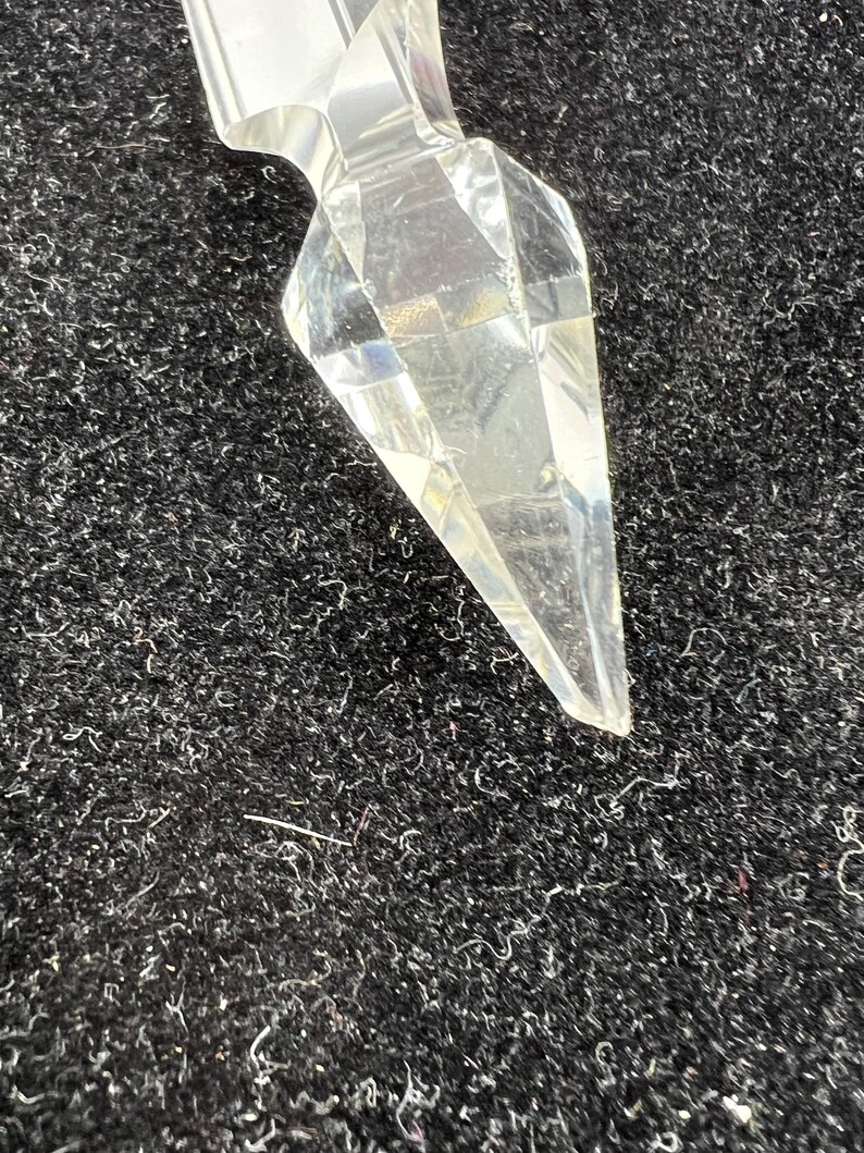 Puede incluir: Un objeto de cristal transparente y facetado con un extremo puntiagudo. El cristal tiene un dise&ntilde;o geom&eacute;trico y refleja la luz, creando un efecto brillante. El objeto est&aacute; sobre un fondo oscuro y texturizado.