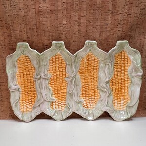 May include: A rectangular ceramic serving platter shaped like four ears of corn arranged side by side. The corn kernels are yellow and orange, while the surrounding husks are textured and painted in shades of light green and cream.