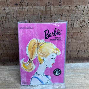 May include: A pink Barbie Teen-Age Fashion Model booklet. The cover features a blonde woman with a ponytail and a blue necklace. The text reads "Barbie" and "Teen-Age Fashion Model". The booklet is a re-creation of the original 1959 booklet.