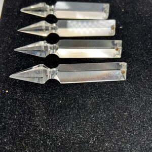 Puede incluir: Cuatro piezas decorativas transparentes con forma de cristal y extremos puntiagudos. Cada pieza tiene un cuerpo rectangular y un peque&ntilde;o agujero redondeado en el extremo. Los objetos est&aacute;n dispuestos sobre una superficie oscura y texturizada.
