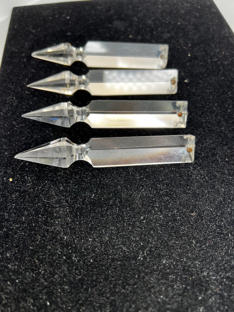 Puede incluir: Cuatro piezas decorativas transparentes con forma de cristal y extremos puntiagudos. Cada pieza tiene un cuerpo rectangular y un peque&ntilde;o agujero redondeado en el extremo. Los objetos est&aacute;n dispuestos sobre una superficie oscura y texturizada.