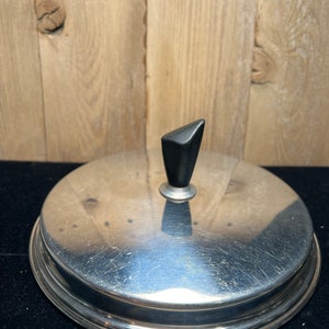 May include: A silver-colored lid with a black knob on top. The lid has a smooth, reflective surface and a raised edge around the perimeter.