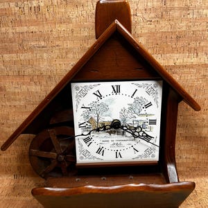 May include: A wooden clock with a house-like frame. The clock face features Roman numerals and a black and white pastoral scene. The clock has a water wheel on the side and the text "HOME TO THANKSGIVING" on the clock face.
