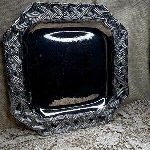 May include: A square, silver-toned serving tray with a woven border design. The tray's interior is a reflective, dark surface. The woven border adds a decorative element to the tray, suitable for serving or display.