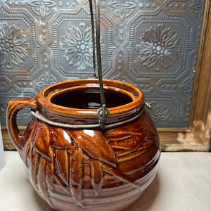 May include: A brown ceramic jar with a metal handle. The jar has a design of leaves and a basket weave pattern.