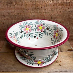 May include: White ceramic colander with a floral design in pink, yellow, and green. The colander has a red rim and sits on a matching base. The design features small holes for draining.