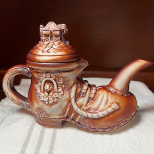 May include: A brown ceramic teapot shaped like a boot with a small house on the lid. The teapot has a white and brown design with a small animal peeking out of a window.