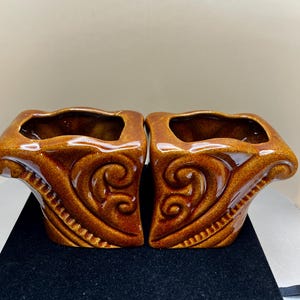 May include: Two matching brown ceramic planters with a glossy finish. Each planter has a square opening and a decorative, curved design on the sides. The planters are placed side by side.