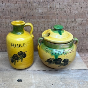 May include: Yellow ceramic oil and mustard containers. The oil container has a handle and the word "HUILE" in black. The mustard container has a lid and the word "MOUTARDE" in black. Both have olive branch illustrations.