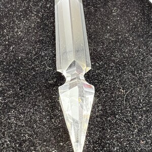 Puede incluir: Un prisma de cristal transparente y facetado con una punta. El cristal tiene un cuerpo rectangular con una parte superior biselada y una parte inferior decorativa y puntiaguda. El cristal se muestra sobre un fondo oscuro y texturizado.