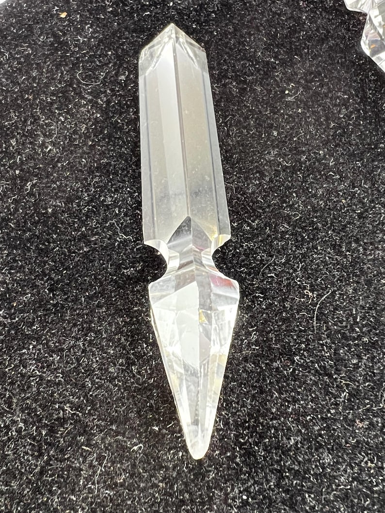 Puede incluir: Un prisma de cristal transparente y facetado con una punta. El cristal tiene un cuerpo rectangular con una parte superior biselada y una parte inferior decorativa y puntiaguda. El cristal se muestra sobre un fondo oscuro y texturizado.