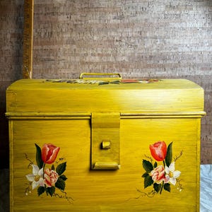 May include: A yellow metal storage box with a hinged lid and a latch. The box is decorated with hand-painted floral designs in shades of red, white, and green.